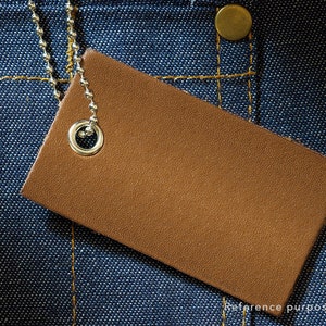 May include: A brown leather tag with a silver metal ring attached to a chain. The tag is on a blue denim background.