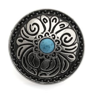 May include: A silver-coloured, round button with an ornate floral design. The button features a central, round, turquoise-coloured stone. The outer edge has a textured, repeating pattern. The button is likely for clothing or accessories.