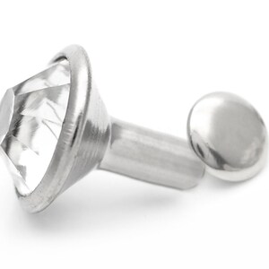 May include: A silver-toned metal rivet with a clear, faceted crystal top. The rivet has a round base and a long, cylindrical shank.