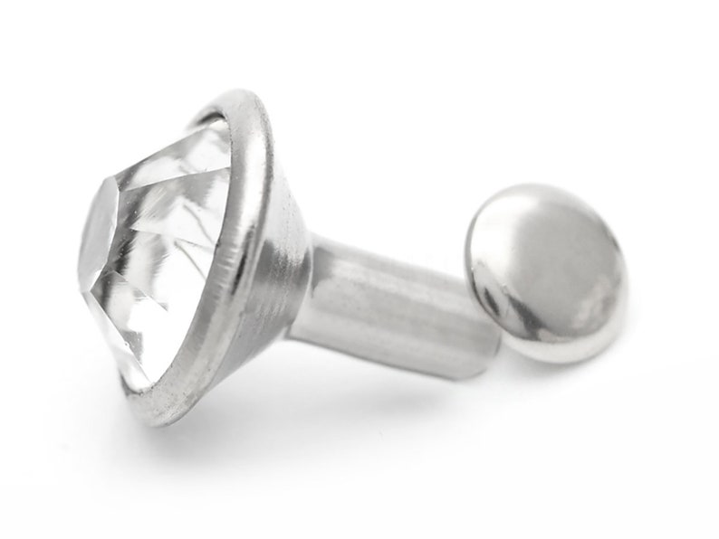 May include: A silver-toned metal rivet with a clear, faceted crystal top. The rivet has a round base and a long, cylindrical shank.