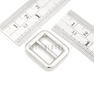 May include: Silver metal buckle measuring 1 inch by 1 inch. The buckle is rectangular with a smooth, polished finish.