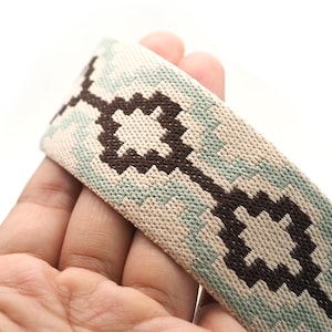K&ouml;nnte beinhalten: Nahaufnahme eines gewebten Riemens mit geometrischem Muster. Der Riemen weist ein sich wiederholendes Design aus dunkelbraunen und hellblauen Formen auf beigem Hintergrund auf. Der Riemen wird in einer Hand vor wei&szlig;em Hintergrund gehalten.
