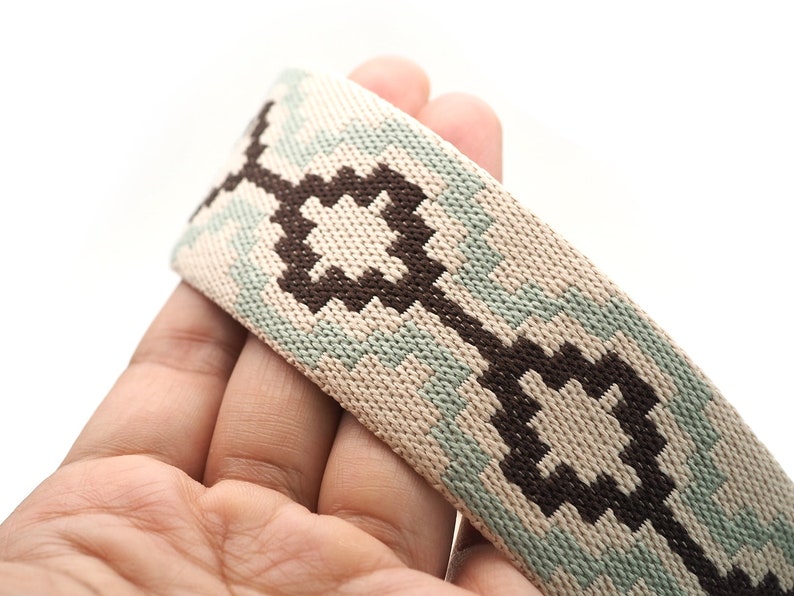 K&ouml;nnte beinhalten: Nahaufnahme eines gewebten Riemens mit geometrischem Muster. Der Riemen weist ein sich wiederholendes Design aus dunkelbraunen und hellblauen Formen auf beigem Hintergrund auf. Der Riemen wird in einer Hand vor wei&szlig;em Hintergrund gehalten.