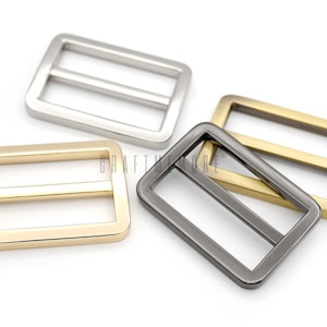 May include: Four metal slider buckles in different finishes: silver, gold, black, and brass. The buckles are rectangular with a center bar.