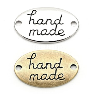 Può includere: Due etichette ovali in metallo con le parole "hand made" scritte in corsivo nero. L'etichetta a sinistra è argentata, mentre quella a destra è dorata.