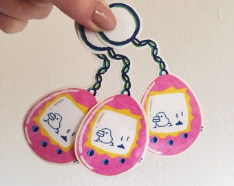 May include: Three pink and white stickers in the shape of a retro handheld game. Each sticker features a cartoon duck character on a screen. The stickers are connected by green and blue chains and held by a hand.