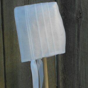 May include: White bonnet with a satin finish and four vertical pleats. The bonnet has white satin ties.