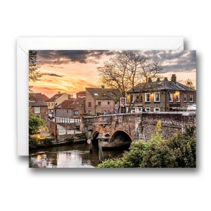 May include: A brick bridge over a river with a pub on the right side of the bridge. The buildings are brick and have a rustic look. The sky is a vibrant orange and pink sunset.