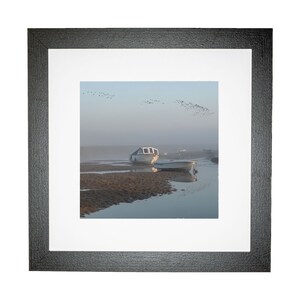 May include: A framed photograph of two white boats on a calm body of water with a flock of birds flying overhead. The water is shallow and the sand is visible in the foreground. The sky is a light blue with a hazy mist.