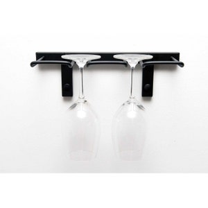 May include: Black metal wine glass rack mounted on a white wall with two clear wine glasses hanging from it.