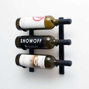 May include: Black metal wall-mounted wine rack with three bottles of wine. The bottles are labeled "Focus", "Showoff", and "Collage van vintage".