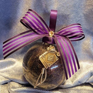 May include: A black glitter Christmas ornament with a gold book charm and a purple and silver striped ribbon bow.