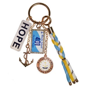 May include: A gold-toned keychain with multiple charms. Charms include a white rectangle with "HOPE" in black, a gold anchor, a blue license plate with "401 Rhode Island Ocean State", and a round charm with a nautical design. The keychain has a braided blue, white, and yellow tassel.