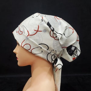 Puede incluir: Una cofia blanca con un patrón de estetoscopio rojo y negro. La cofia tiene una corbata que se puede atar en la parte posterior del cuello.