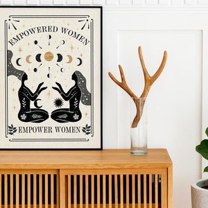 May include: A black and white poster with a beige background featuring two women with their arms raised, surrounded by stars, moons, and a sun. The text "EMPOWERED WOMEN" is at the top of the poster and "EMPOWER WOMEN" is at the bottom.