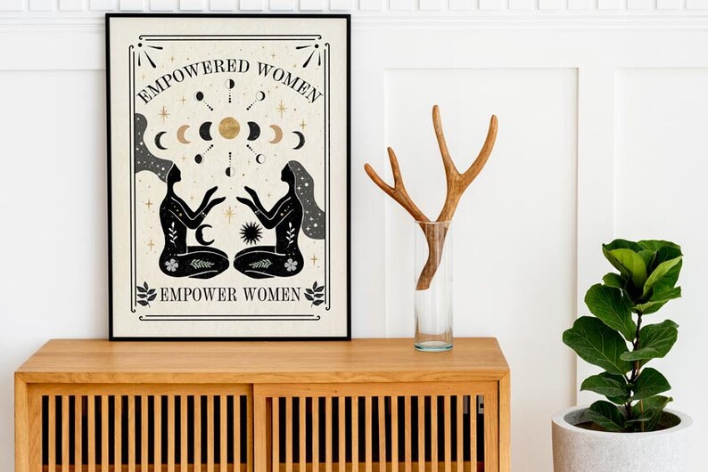 May include: A black and white poster with a beige background featuring two women with their arms raised, surrounded by stars, moons, and a sun. The text "EMPOWERED WOMEN" is at the top of the poster and "EMPOWER WOMEN" is at the bottom.