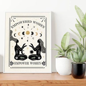 May include: A beige printable art print with black and gold illustrations of two women facing each other. The women are surrounded by moons and stars. The text "EMPOWERED WOMEN EMPOWER WOMEN" is printed in black at the top and bottom of the print.