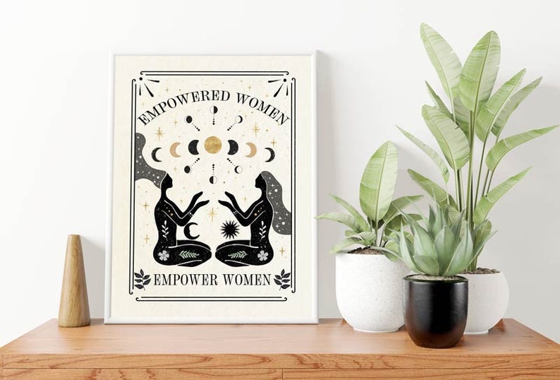 May include: A beige printable art print with black and gold illustrations of two women facing each other. The women are surrounded by moons and stars. The text "EMPOWERED WOMEN EMPOWER WOMEN" is printed in black at the top and bottom of the print.