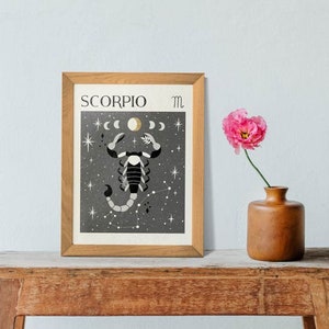 May include: A framed print with a black and white illustration of a scorpion with a crescent moon and stars on a dark gray background. The text "SCORPIO" and the astrological symbol for Scorpio are printed in the top left and right corners of the print.