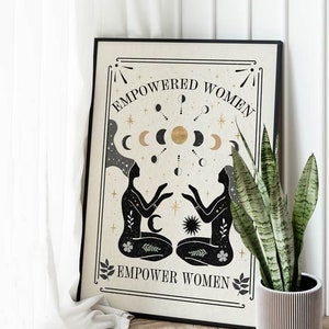 May include: A beige poster with black text that reads "Empowered Women Empower Women". The poster features a black and gold illustration of two women sitting back to back with their hands raised. The illustration is surrounded by a circle of moons and stars.