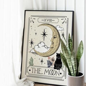 K&ouml;nnte beinhalten: Ein schwarzer gerahmter Druck einer Tarotkarte, die die Mondkarte darstellt. Die Karte zeigt einen Halbmond mit einem Gesicht, eine schwarze Katze und Sterne. Der Text "XVIII. THE MOON." ist auf der Karte gedruckt.