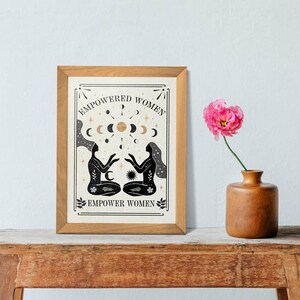 May include: A framed print with a black and white illustration of two women sitting back to back with their hands raised. The illustration is surrounded by a gold moon and star design. The text "EMPOWERED WOMEN" is at the top of the print and "EMPOWER WOMEN" is at the bottom.