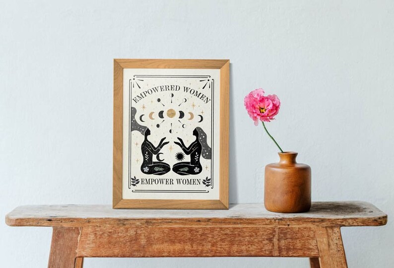 May include: A framed print with a black and white illustration of two women sitting back to back with their hands raised. The illustration is surrounded by a gold moon and star design. The text "EMPOWERED WOMEN" is at the top of the print and "EMPOWER WOMEN" is at the bottom.