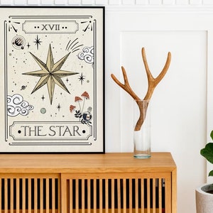 May include: A framed print of a tarot card depicting The Star card. The card features a large, eight-pointed star with a beige background and black outlines. The card is framed in black and is sitting on a wooden cabinet.