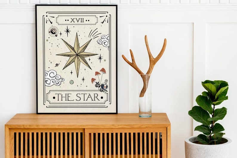 May include: A framed print of a tarot card depicting The Star card. The card features a large, eight-pointed star with a beige background and black outlines. The card is framed in black and is sitting on a wooden cabinet.