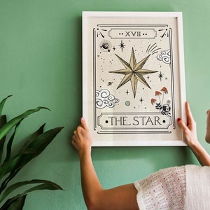 May include: A white framed print of a tarot card depicting The Star card. The card features a large, gold star with a white outline and a white background. The card also features a variety of other symbols, including clouds, stars, and mushrooms.