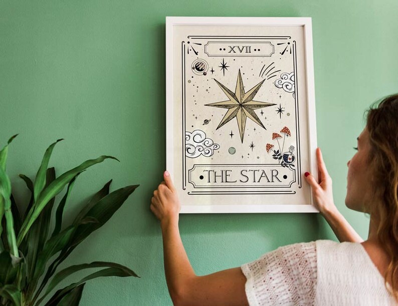 May include: A white framed print of a tarot card depicting The Star card. The card features a large, gold star with a white outline and a white background. The card also features a variety of other symbols, including clouds, stars, and mushrooms.