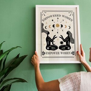 May include: A white framed print with a black and gold design. The print features two stylized figures with their arms outstretched towards each other. The text "EMPOWERED WOMEN" and "EMPOWER WOMEN" is printed in black on the print.