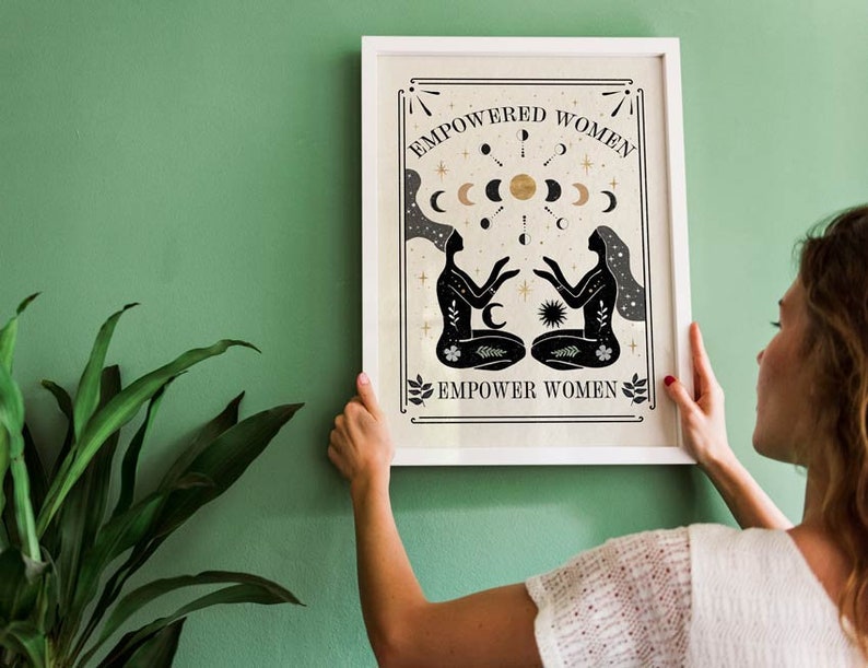 May include: A white framed print with a black and gold design. The print features two stylized figures with their arms outstretched towards each other. The text "EMPOWERED WOMEN" and "EMPOWER WOMEN" is printed in black on the print.