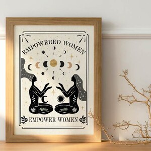 May include: A framed print with a beige background and black silhouettes of two women facing each other. The text "EMPOWERED WOMEN" is at the top and "EMPOWER WOMEN" is at the bottom. The print features a gold sun and moons.
