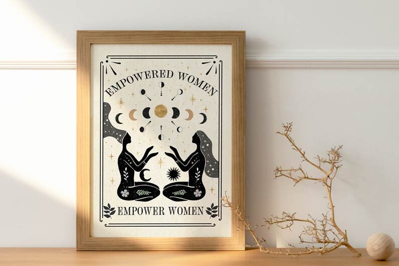 May include: A framed print with a beige background and black silhouettes of two women facing each other. The text "EMPOWERED WOMEN" is at the top and "EMPOWER WOMEN" is at the bottom. The print features a gold sun and moons.