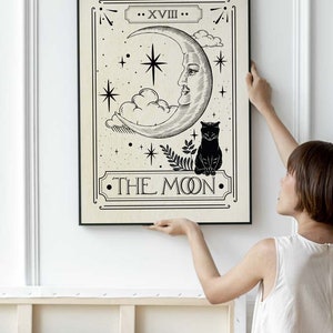 May include: A black and white tarot card illustration of the Moon card. The card features a crescent moon with a human face, a black cat, and stars. The text "XVIII THE MOON" is printed on the card.