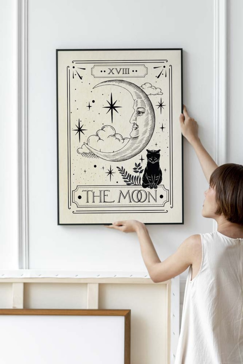 May include: A black and white tarot card illustration of the Moon card. The card features a crescent moon with a human face, a black cat, and stars. The text "XVIII THE MOON" is printed on the card.