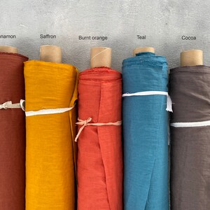 May include: Five rolls of fabric in different colors: cinnamon, saffron, burnt orange, teal, and cocoa. The fabric is rolled up and tied with a white ribbon.