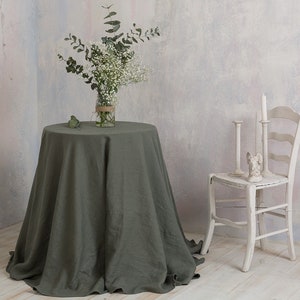 May include: A green linen tablecloth draped over a round table with a vase of white flowers and greenery. A white wooden chair with candlesticks sits to the right of the table.