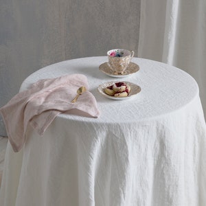 May include: A white linen tablecloth with a pink linen napkin, a gold spoon, a teacup and saucer with floral pattern, and a plate with three cookies on a round table.