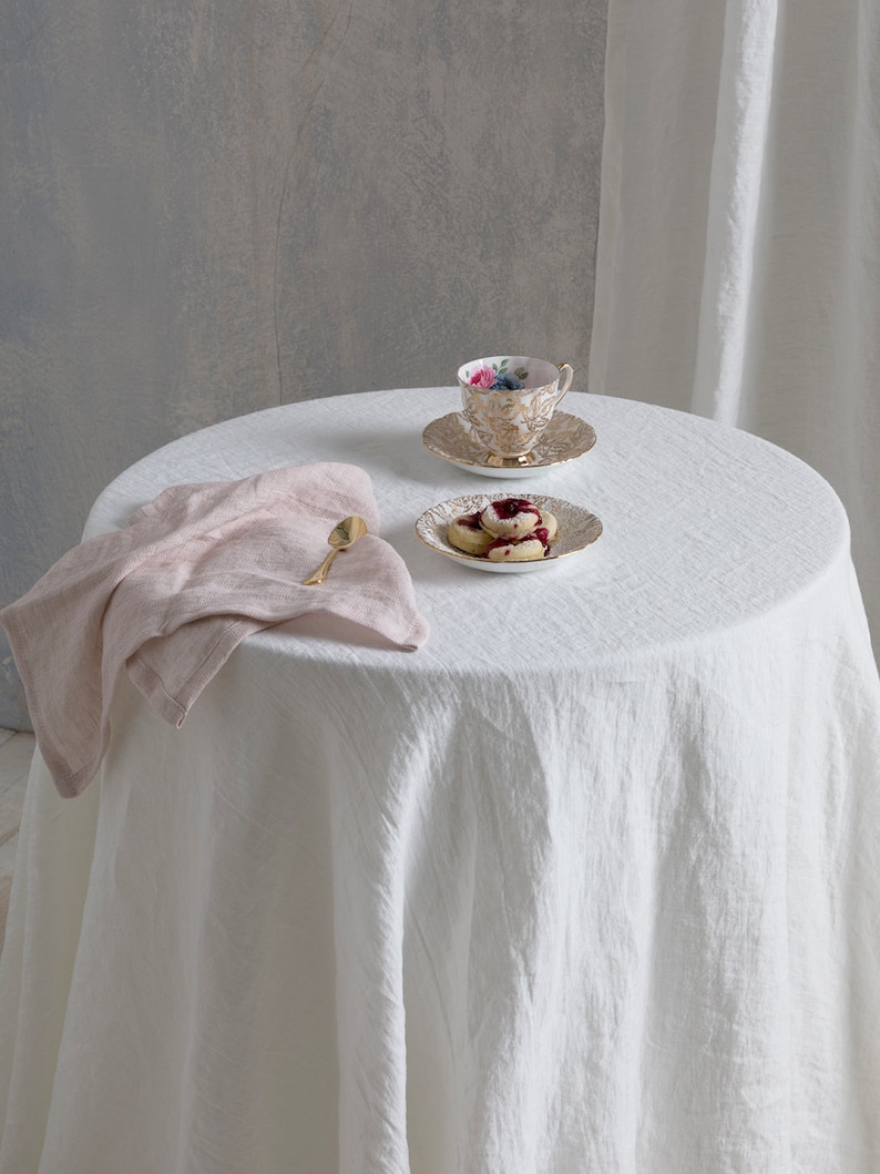 May include: A white linen tablecloth with a pink linen napkin, a gold spoon, a teacup and saucer with floral pattern, and a plate with three cookies on a round table.