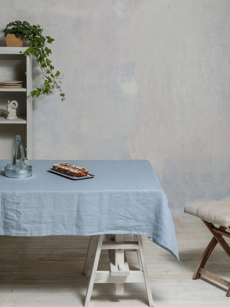 May include: A light blue linen tablecloth draped over a white wooden table with a loaf of bread on a plate. The table is set against a white wall with a potted plant in the background.