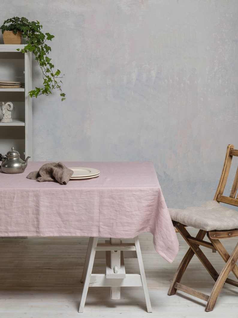 May include: A pink linen tablecloth draped over a white table with a wooden chair and a potted plant in the background.