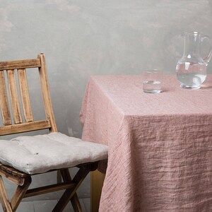 May include: A wooden chair with a light grey cushion sits next to a table covered in a pink linen tablecloth. A clear glass jug and a glass of water are on the table.