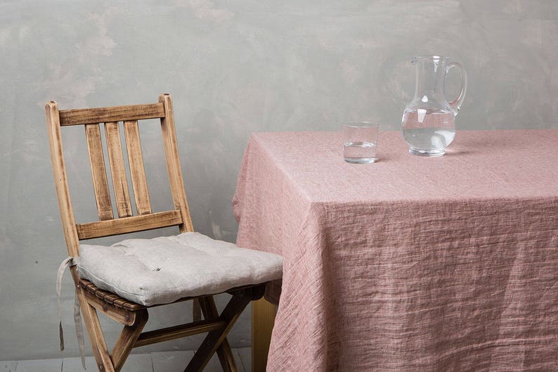 May include: A wooden chair with a light grey cushion sits next to a table covered in a pink linen tablecloth. A clear glass jug and a glass of water are on the table.