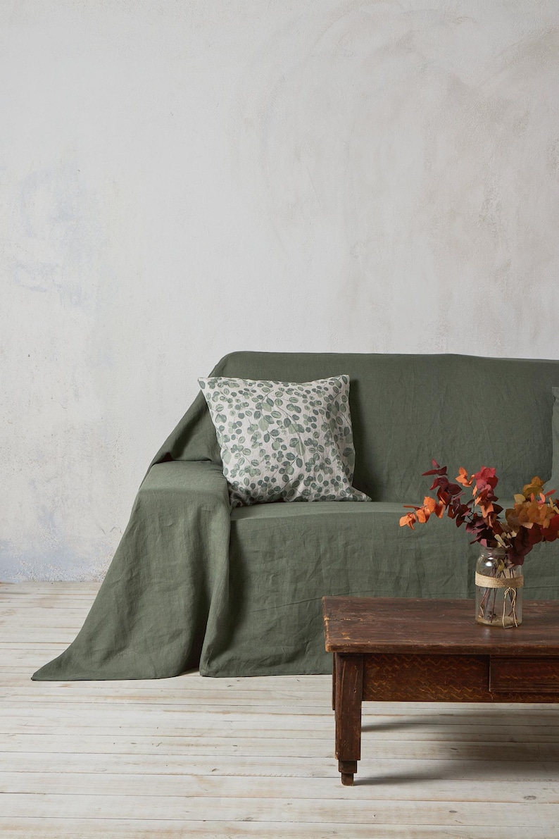 Op de afbeelding: Een groene linnen sofa deken met een gepatroneerd kussen en een donkerhouten salontafel. Een vaas met gedroogde bloemen staat op de tafel.