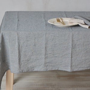 May include: A gray and white striped linen tablecloth with a white plate, two gold forks, and a white napkin on top. The tablecloth is draped over a wooden table.