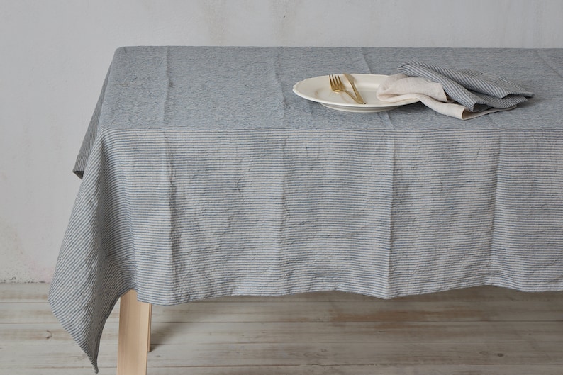 May include: A gray and white striped linen tablecloth with a white plate, two gold forks, and a white napkin on top. The tablecloth is draped over a wooden table.