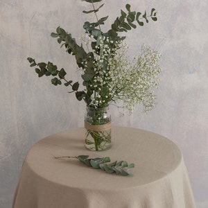 Puede incluir: Un ramo de aliento de beb&eacute; blanco y eucalipto verde en un frasco de vidrio transparente atado con cuerda, sentado sobre un mantel de lino beige.