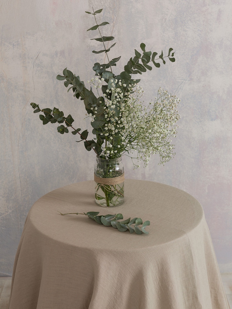 Puede incluir: Un ramo de aliento de beb&eacute; blanco y eucalipto verde en un frasco de vidrio transparente atado con cuerda, sentado sobre un mantel de lino beige.